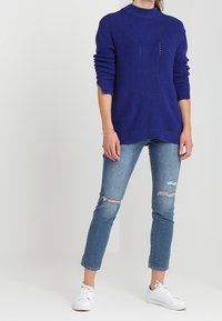 Modrý pletený svetr s vysokým límcem, který má strukturované žebrování a vzorové detaily. Spárován se světlými modrými roztrhanými džíny a bílými teniskami.