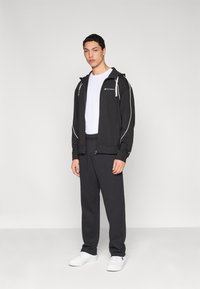 Sweat à capuche noir zippé avec des accents blancs, assorti à un pantalon de survêtement noir et des baskets blanches. La tenue présente un design simple et décontracté.