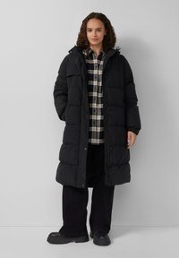 Duga crna puffer jakna s kapuljačom, s patentnim zatvaračem i dugmadi, nosi se preko karirane košulje i crnih hlača.