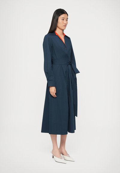 Granatowa sukienka kopertowa z długimi rękawami, zapinana na guziki z przodu i z paskiem w talii. Posiada kontrastujący pomarańczowy kołnierz oraz gładką fakturę.