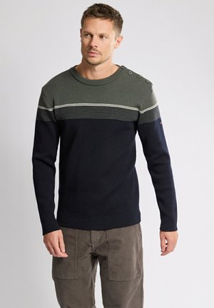 Homme portant un pull coloré à bloc, avec une partie supérieure verte et une partie inférieure bleu marine, des boutons sur l'épaule, et un pantalon marron, se tenant devant un fond uni.