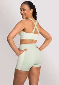 Top sportivo verde chiaro con spalline incrociate, abbinato a pantaloni corti a vita alta. Texture liscia, design aderente e dettagli di marca minimi.