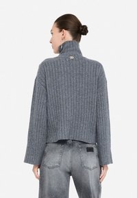 Maglione grigio a coste con colletto alto e design cropped, caratterizzato da maniche lunghe e un piccolo logo sul retro. Abbinato a jeans a vita alta.