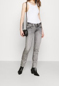 Donna che indossa un top senza maniche bianco, jeans slim-fit grigi, stivali neri alla caviglia e porta una piccola borsa a tracolla nera.