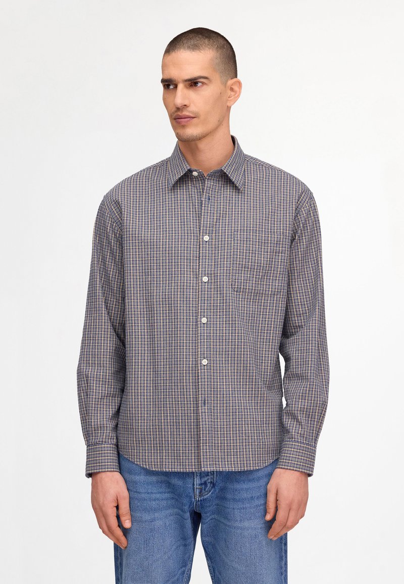 Homme aux cheveux courts portant une chemise boutonnée à manches longues à carreaux gris et bleus, ainsi qu'un jean bleu, debout devant un fond blanc uni.