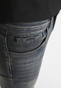 Tamno plave traperice uskog kroja, s blagim izblijedjenjem, oznakom na stražnjem džepu i klasičnim detaljima šivanja.