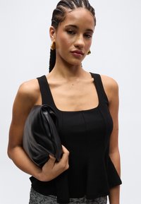 Γυναίκα με πλεγμένα μαλλιά φοράει μαύρο μπλουζάκι χωρίς μανίκια και χρυσούς σκουλαρίκια, κρατώντας μια μαύρη δερμάτινη τσάντα μπροστά σε απλή φόντο.