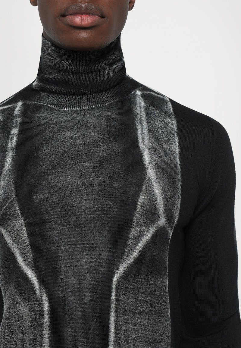 Primo piano di una persona che indossa un maglione a collo alto nero e grigio con un design geometrico e un colletto a coste su uno sfondo neutro.