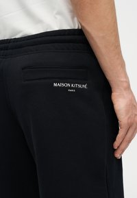 Primer plano de una persona que lleva pantalones deportivos negros de Maison Kitsuné con cintura elástica y texto de logo blanco en la zona del bolsillo trasero.