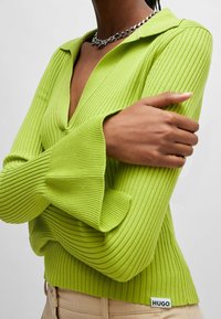 Sweter w limonkowym kolorze, prążkowany, z kołnierzykiem, głębokim dekoltem w serek i rękawami dzwonowymi. Posiada małą czarną metkę z napisem "HUGO" na dolnym brzegu.