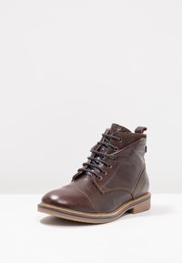 Pánské tmavě hnědé kožené kotníkové boty s černými tkaničkami, s podrobným prošíváním a světlou hnědou podrážkou, umístěné na bílé ploše.