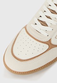 Sapatilhas em creme e castanho com uma caixa de dedos perfurada, materiais de camurça e couro, sola de borracha texturizada e atacadores brancos.
