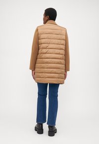 Cappotto imbottito beige con finitura liscia e forma leggermente oversize, abbinato a jeans blu a zampa e stivali neri chunky.