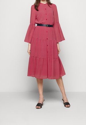 Kobieta w czerwonej, wzorzystej sukience midi z rozszerzanymi rękawami, z przodu na guziki, z czarnym paskiem i czarnymi sandałami na obcasie z otwartymi palcami.