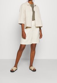 Person iført en beige skjorte med knapper, en olivengrøn t-shirt, beige knælange shorts og beige krydssandaler mod en ensfarvet baggrund.