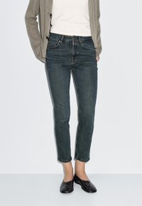 Dark wash denim jeans with a slim fit and slight ankle taper. Worn with a white top and textured gray cardigan. Black slip-on shoes.