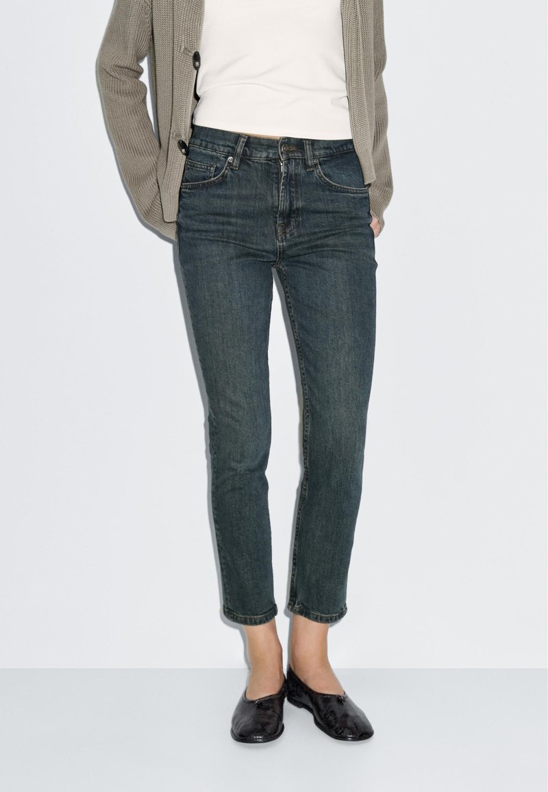 Dark wash denim jeans with a slim fit and slight ankle taper. Worn with a white top and textured gray cardigan. Black slip-on shoes.