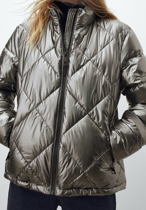 Kobieta ubrana w metalicznie srebrną pikowaną kurtkę puchową z przednim zamkiem i rękami w kieszeniach, w połączeniu z ciemnymi spodniami.
