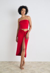 Top corto rojo con tirantes finos combinado con una falda larga, anudada y de gran abertura. Textura de tela suave, diseño minimalista y color vibrante.