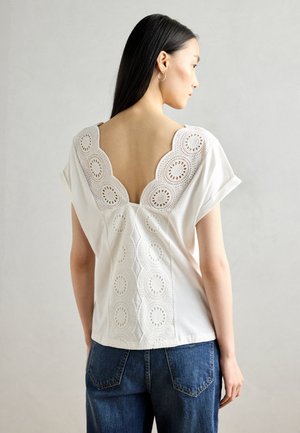 White blouse featuring a deep V-back with intricate lace detailing, short sleeves, and a smooth texture, paired with blue jeans.