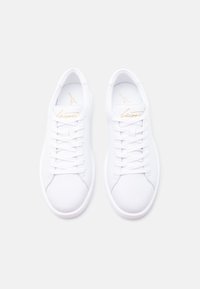 Ζευγάρι λευκά χαμηλά αθλητικά παπούτσια με λευκά κορδόνια και χρυσό λογότυπο με γραφή στη γλώσσα, φωτογραφημένα από πάνω σε λευκό φόντο.