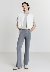 Gilet matelassé blanc avec poches sur un t-shirt gris, associé à un pantalon évasé en tricot gris et des baskets blanches avec des accents beiges.