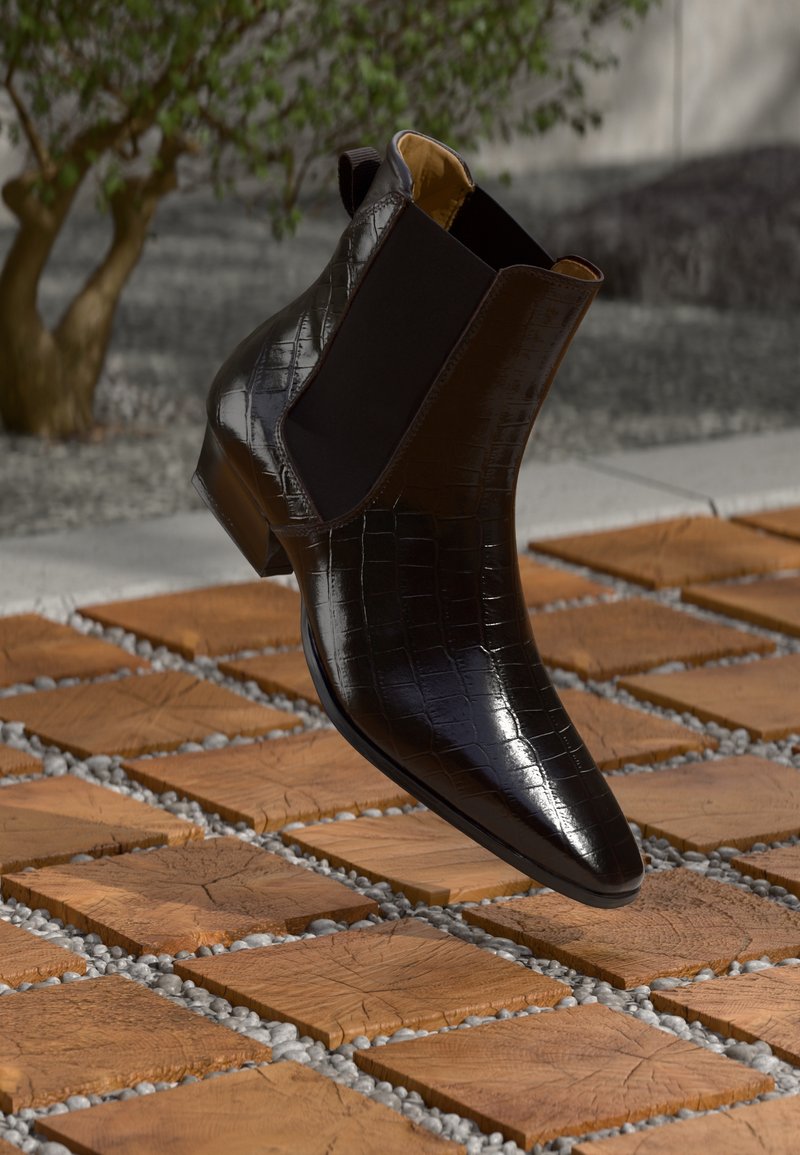 Käekaitsmed mustast läikivast nahast, krokodillimustriga, hõljumas väljas puidust ruudukujuliste plaatide kohal, mille vahel on väikesed hallid kivid.