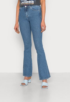 Femme portant un jean évasé taille haute bleu clair avec des fentes sur les côtés et des sandales à talons blocs bleu clair à bouts ouverts, debout sur fond blanc.