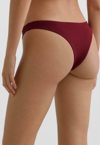 Spodní díl bikin v texturované bordó látce s nízkým střihem, vyznačující se minimalistickým designem a hladkým povrchem.