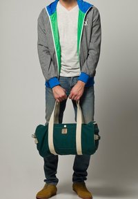 Grön kanvas resväska med krämfärgade remmar, rund form, metallbeslag och liten logotyp-logga. Modellen håller väskan med båda händerna.