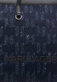 Denimová taška přes rameno s tmavomodrou tkaninou a texturovaným vzorem. Černé logo a akcenty. Obsahuje detail kovového uchycení popruhu.