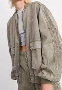 Olive green bomber jacket with ribbed cuffs, two front pockets, and subtle three-stripe detailing on the sleeves. Cotton blend material.