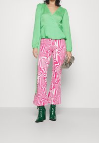 Top verde a maniche lunghe plissettato, pantaloni a palazzo rosa e bianchi con swirls, e stivali alla caviglia verdi lucidi, con una piccola borsa a tracolla argentata con borchie.