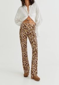 Kobieta w wzorzystych spodniach z wysokim stanem i wiązaniami, luźnej białej koszuli na guziki oraz brązowych butach na platformie, stojąca na tle białym.