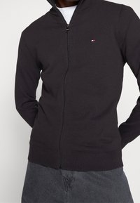 Tmavě šedý zip-up cardigan s vysokým límcem, žebrovanými manžetami a lemem. Obsahuje malé logo s červenými a bílými akcenty na prsou.