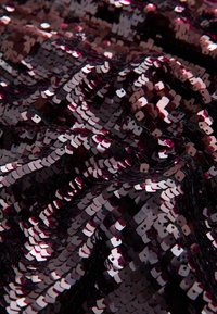 Tumepruun litritekangas tekstureeritud pinnaga. Litrid on paigutatud kattuvatesse ridadesse, mis peegeldavad valgust ja loovad sätendava efekti.