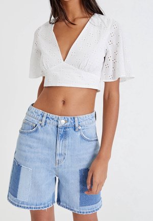 Top corto blanco con patrón de ojal y mangas cortas, combinado con pantalones cortos de mezclilla azul claro con acentos de parches y dobladillos deshilachados.