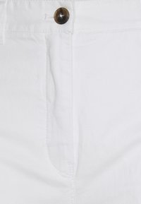 Gros plan de la taille d'un pantalon blanc avec un seul bouton noir et des coutures visibles autour de l'ouverture du bouton et de la braguette.