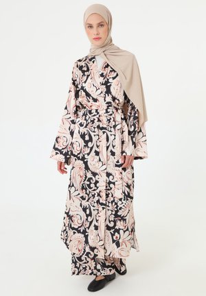 Kvinde iført en beige hijab og en lang sort og cremefarvet blomsterpræget kjole med bindebælte, stående mod en ensfarvet hvid baggrund.