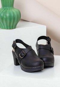 Zwarte leren klompen met een stevige hak, voorzien van een verstelbare band en zilveren gespen. De zool heeft contrasterende stiksels en textuur.