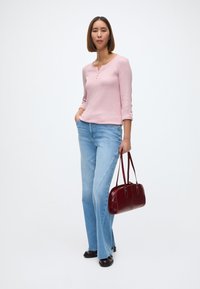 Ροζ ανοιχτό μπλουζάκι με μακριά μανίκια και τρία κουμπιά, συνδυασμένο με ανοιχτό μπλε φαρδύ τζιν. Κρατά μια μπορντό τσάντα και φοράει μαύρα παπούτσια.