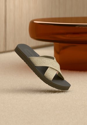 Beige sandaal met gekruiste riemen van gestructureerde stof en een donkere zool, liggend op een beige tapijt naast een bruine afgeronde tafel.