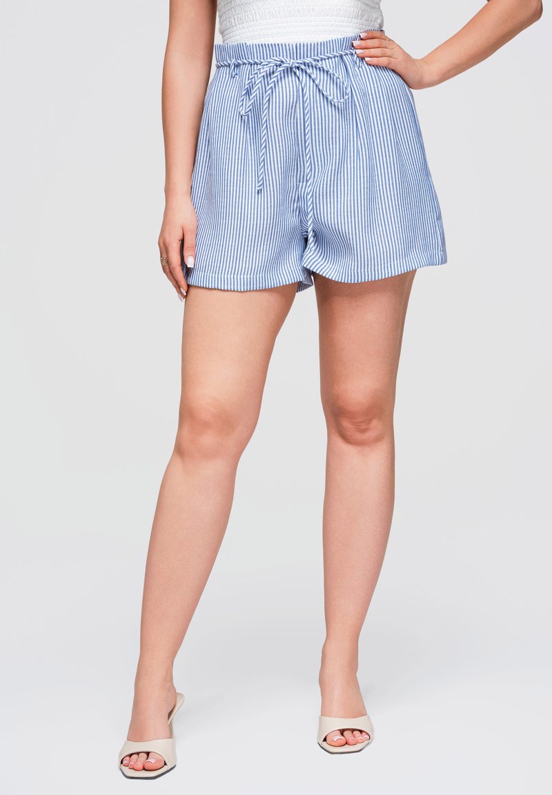 Femme portant un short rayé vertical bleu et blanc avec une ceinture nouée à la taille et des talons blancs ouverts à l'avant, debout avec une main sur la hanche.