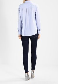 Frau trägt ein hellblau gestreiftes Hemd mit Knöpfen, dunkle enge Jeans und silberne High Heels, steht mit dem Rücken zu einer weißen Wand.