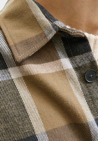 Camicia di flanella con motivo a quadri marrone, nero e bianco, presenta colletto, tessuto testurizzato e dettaglio del bottone nero.