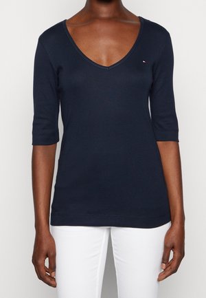 Ciemnogranatowa koszulka z dekoltem w serek i rękawami do łokcia, noszona przez osobę o średnio ciemnej skórze, zestawiona z białymi spodniami, na jednolitym tle.