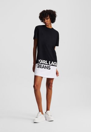 Robe t-shirt noir et blanc featuring un col rond, des manches courtes, et un texte en gras "KARL LAGER JEANS" à l'ourlet. Texture en coton lisse.