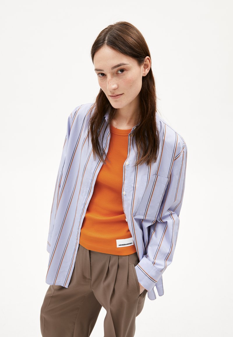 Gestreepte lichtblauwe overhemd met knopjes over een oranje tanktop, gedragen met losse bruine broeken, met een klein zakje en labeldetails.