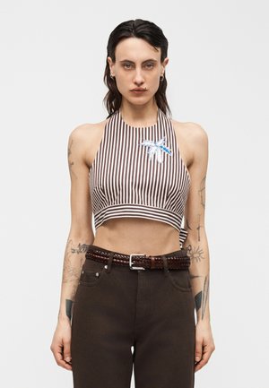 Femme aux cheveux foncés portant un crop top à rayures marron et blanc avec un patch bleu représentant un palmier, et un pantalon foncé avec une ceinture tressée.