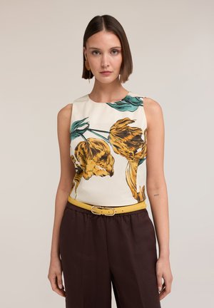 Kvinde med kort brunt hår iført ærmeløs top med stort gult blomsterprint, brune bukser og gul bælte, står mod en ensfarvet baggrund.